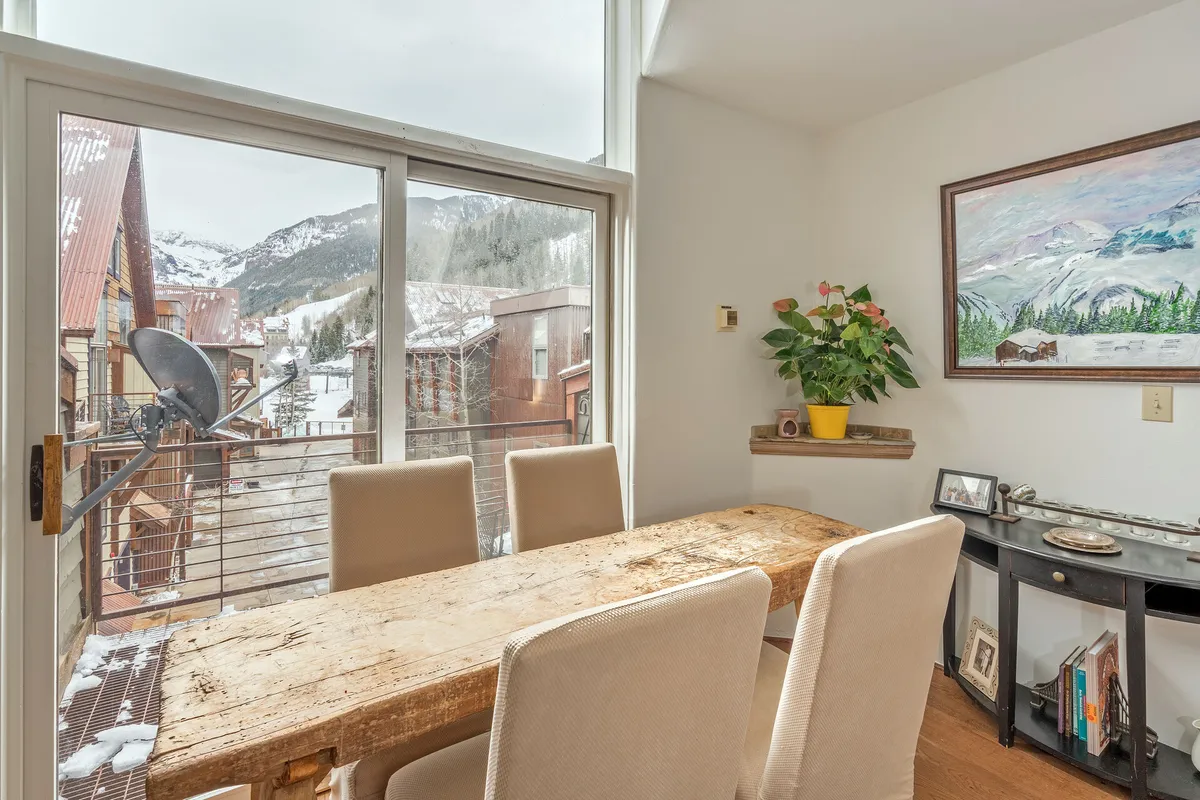 A rustic wooden dining table is set beside large windows framing a stunning mountain view, with touches of greenery creating a warm, homey atmosphere. The scene suggests shared meals, laughter, and memorable moments against a scenic backdrop.