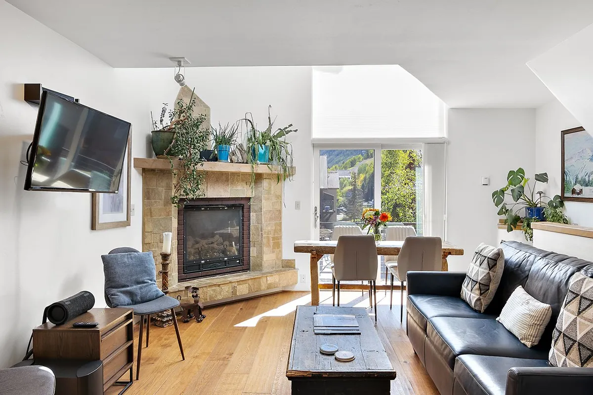 A cozy, sunlit interior featuring a fireplace, a plush sofa, and a dining table set beside large windows with plants. The scene radiates warmth and togetherness, inviting guests to relax, dine, and share stories in a welcoming living room setting.