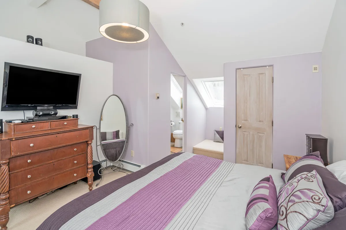 This image features a serene bedroom with a neatly made bed adorned with decorative pillows, a wooden dresser, a television, and a full-length mirror. A doorway leads to an adjacent bathroom, suggesting en suite convenience. Natural light fills the space, contributing to a calm and welcoming atmosphere ideal for rest and relaxation.