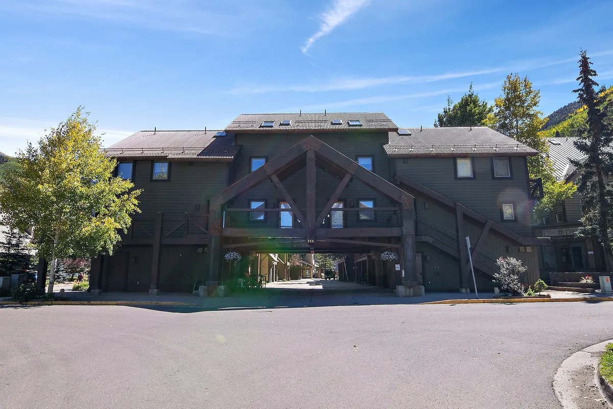 An exterior image highlighting a large wooden structure as the main entrance, surrounded by trees and a welcoming porch area. The scene suggests a space for socializing and relaxing outdoors, with easy access to neighborhood pathways.