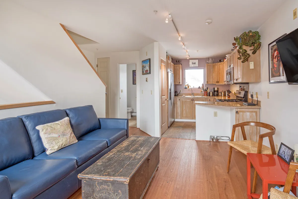 The image showcases the inviting open-concept living area of a Telluride townhouse, featuring a plush blue sofa, a rustic wooden coffee table, and a modern kitchen in the background. A sleek staircase adds architectural interest, while the setup exudes warmth and comfort, ideal for relaxing, socializing, and enjoying cozy gatherings after a day on the slopes.