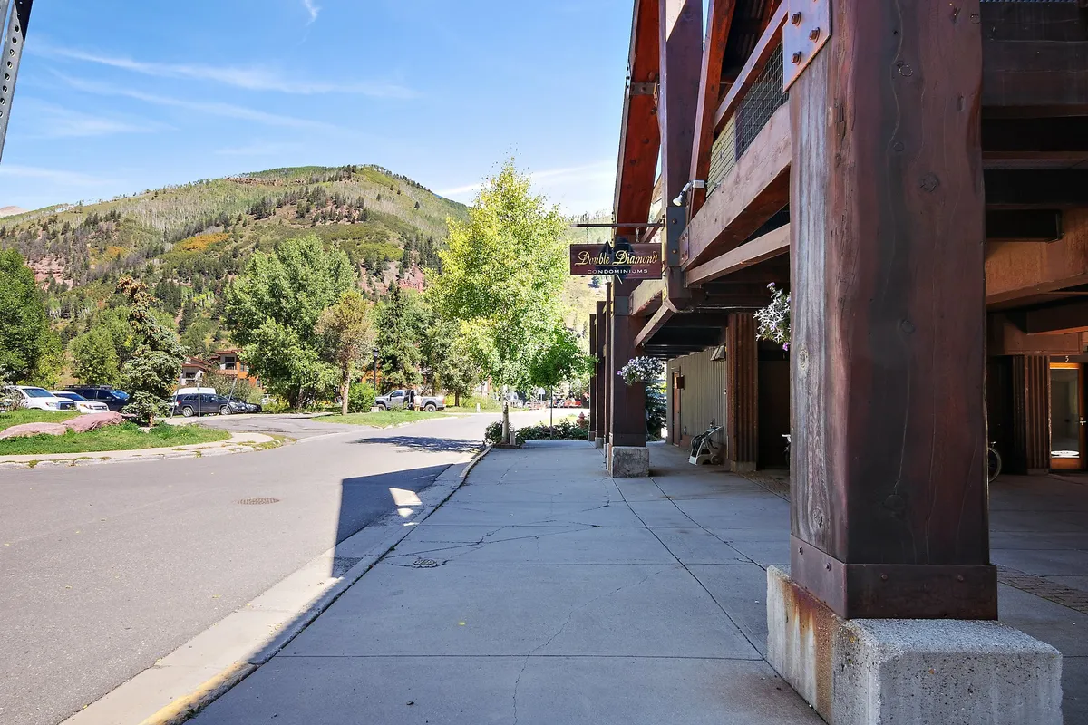 This exterior image features a mountain backdrop, a tree-lined street, and architectural details of the building. The scene exudes a relaxed yet adventurous atmosphere, with clear paths for walking or biking, and the surrounding landscape showing off Telluride’s natural beauty.
