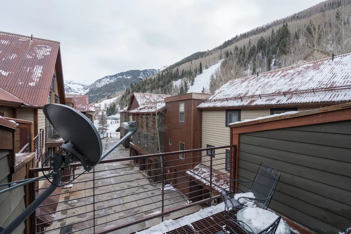 A tranquil winter exterior showing snow-covered mountains, wooden cabins, a metal patio railing, and a cozy patio area. The inviting atmosphere suggests relaxing after a day of skiing while enjoying mountain views and the peaceful snowy landscape.