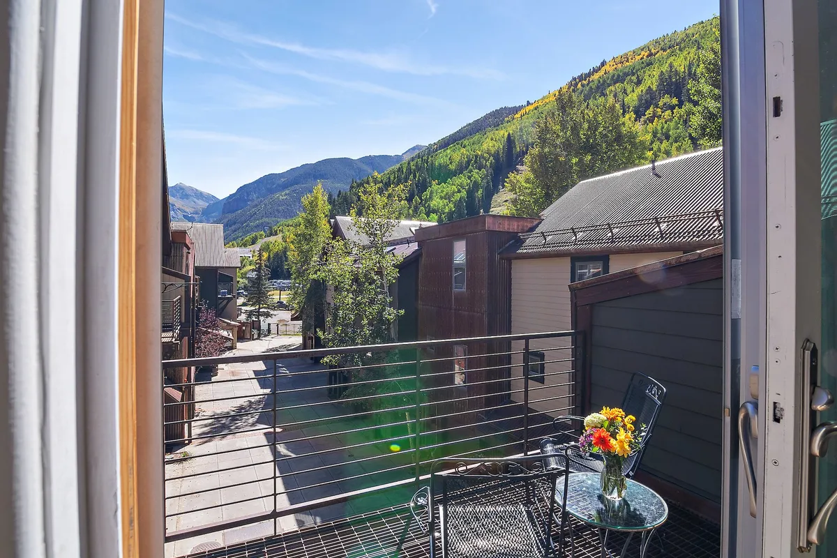 A private balcony adorned with colorful flowers and outdoor seating, set against a sweeping backdrop of Telluride's mountains. Neighboring homes and a clear sky enhance the sense of both privacy and expansive views. The scene suggests a peaceful retreat perfect for morning coffee or evening relaxation.